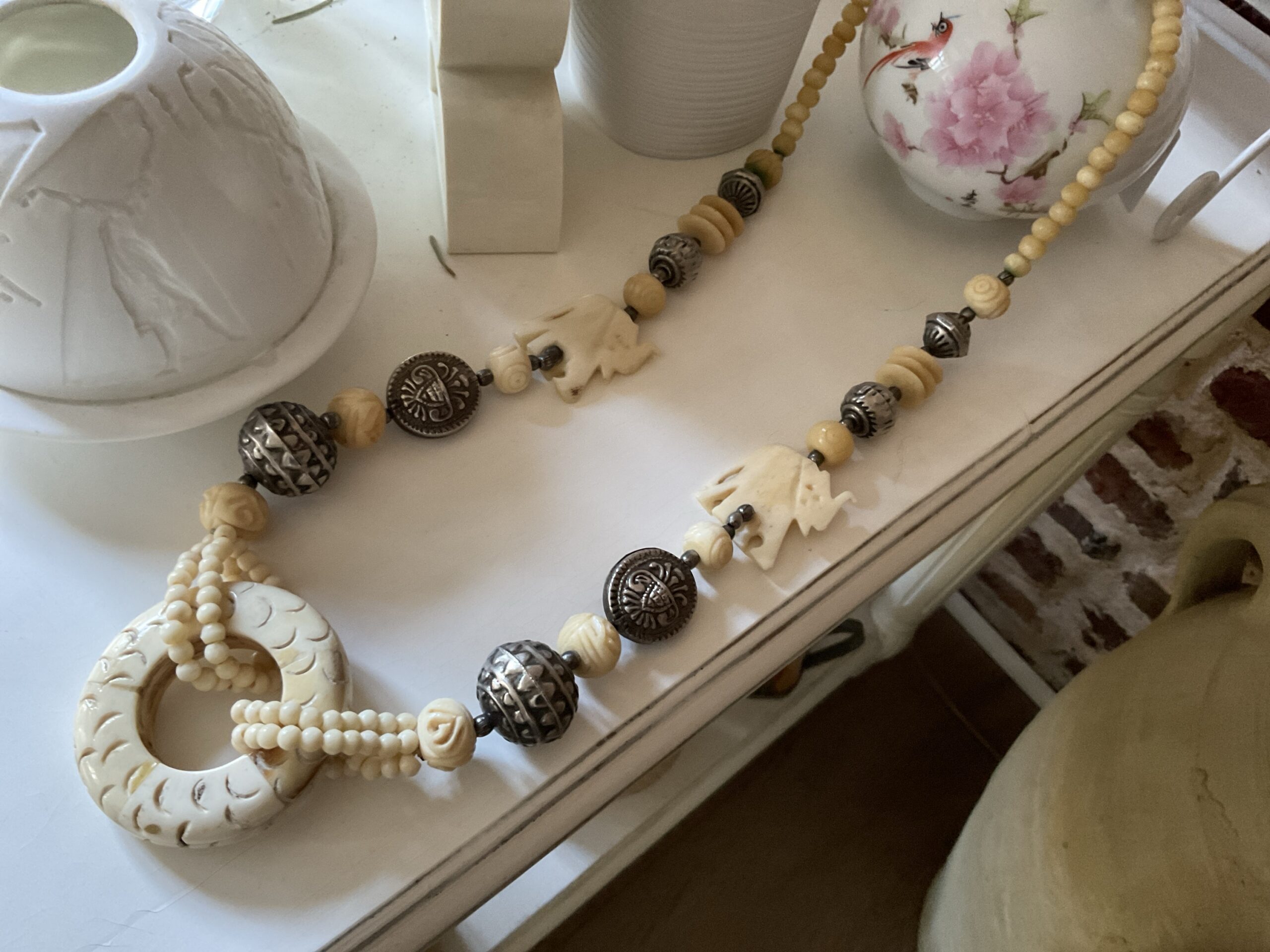 Ce collier est une traversée. Une ligne qui relie les origines à aujourd’hui. Un bijou qui ne se contente pas d’être porté — il s’incarne. Le Cercle des Origines s’articule autour d’un pendentif central sculpté, forme pleine, presque rituelle. Autour de lui, des perles, des volumes, des éléments gravés. Certains évoquent des symboles. D’autres des fragments de mémoire.